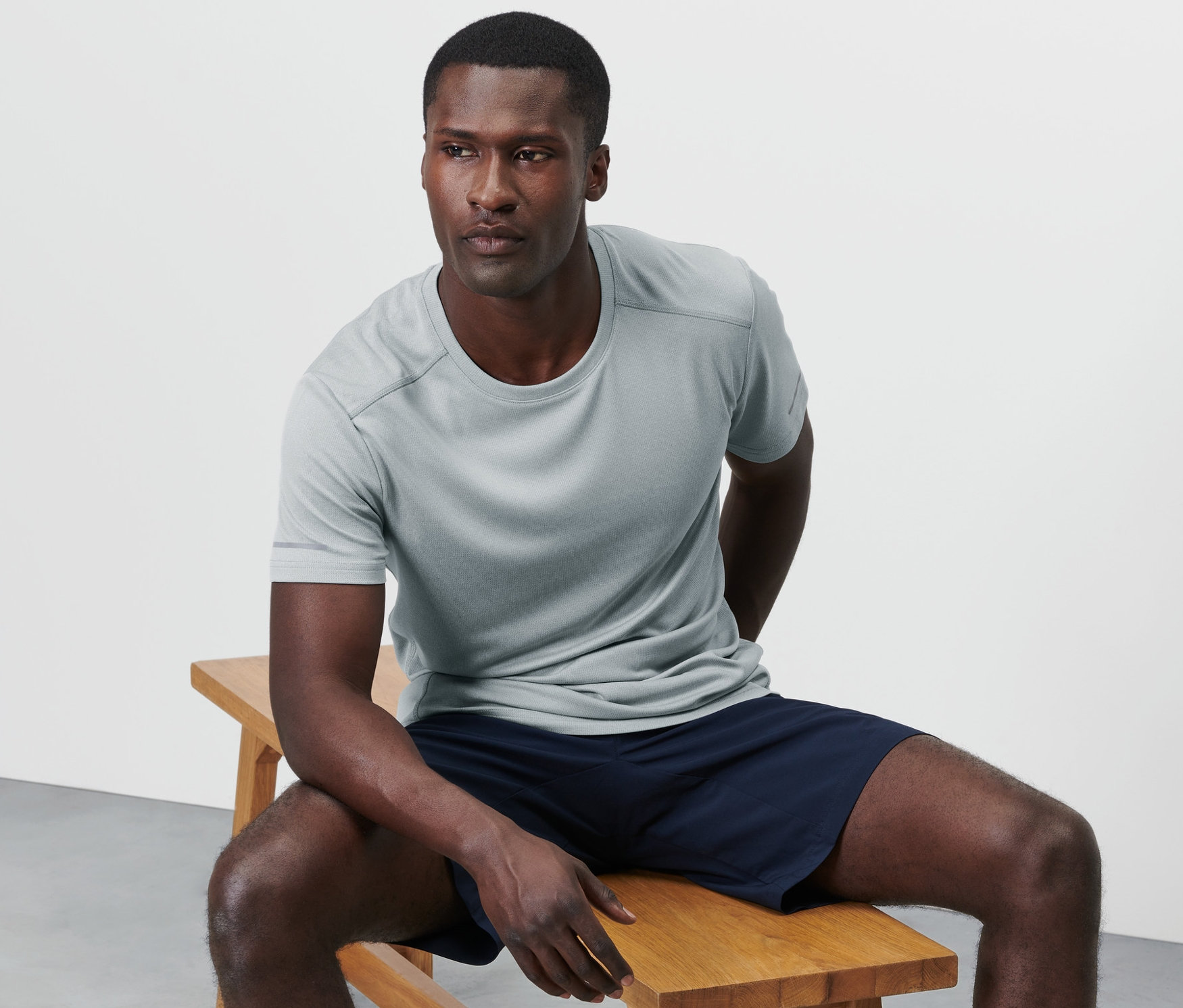 Un homme est assis sur un banc en bois et porte un t-shirt fonctionnel gris chiné.