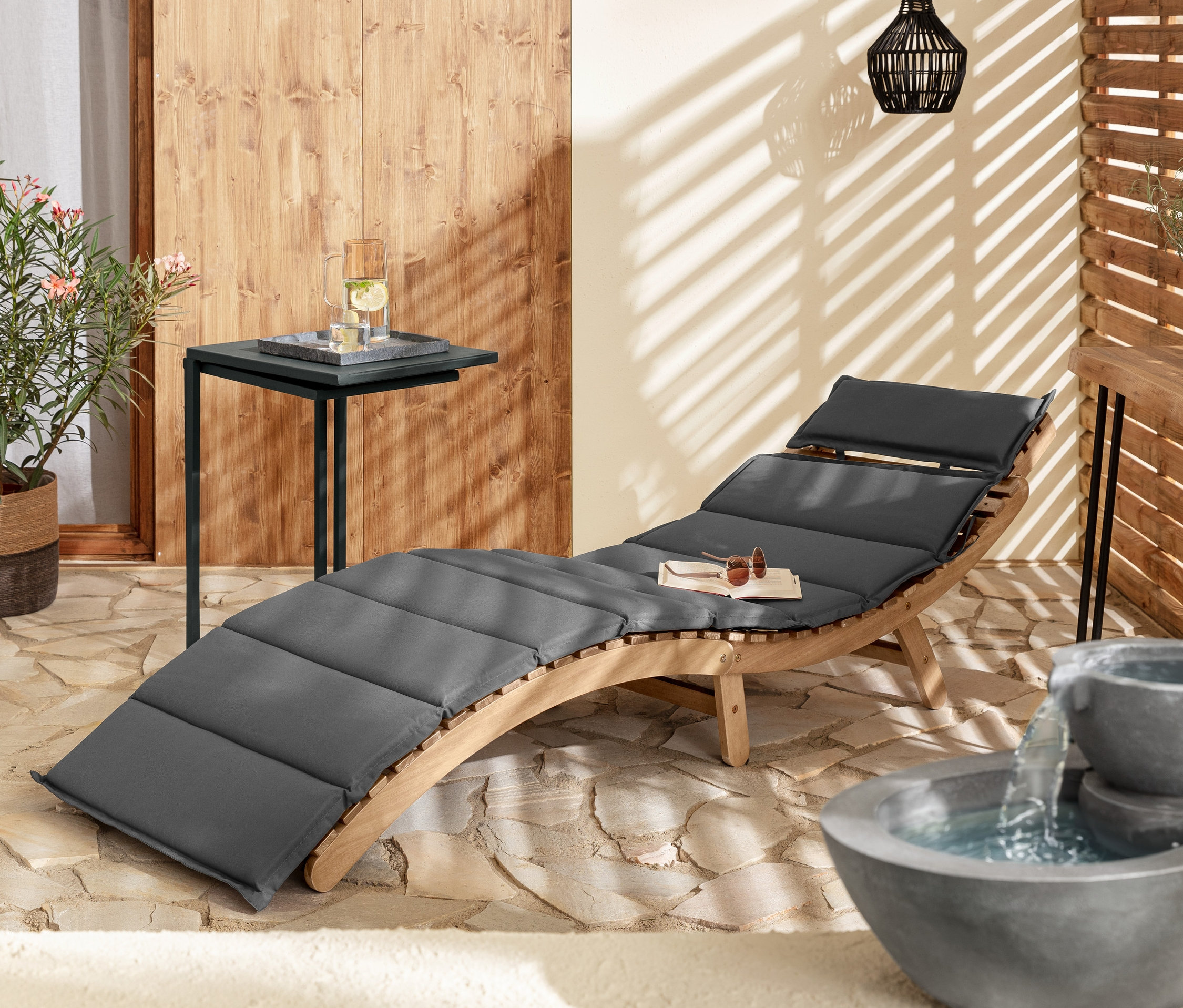 Une chaise longue ondulée et pliable « Lenja » grise est à côté de 2 tables en C et d'une table-console en planches de bois naturel. À l'arrière-plan, il y a une lampe de suspension solaire avec cordon textile et une fontaine solaire de jardin.