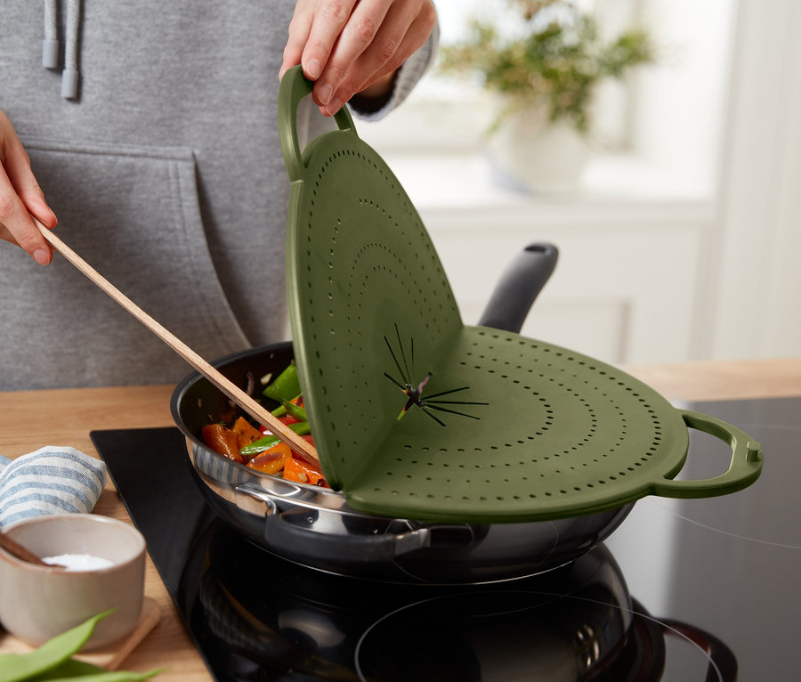 Une personne soulève un couvercle anti-éclaboussures 4 en 1 vert au-dessus d'une poêle avec des légumes.