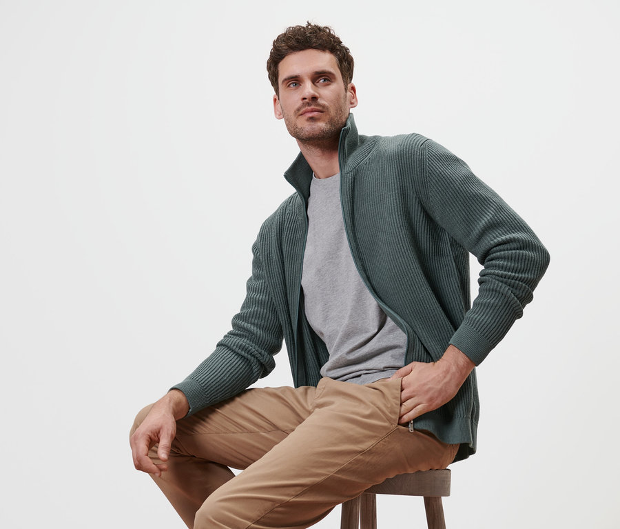 Un homme est assis sur un tabouret et porte une jaquette en maille sauge.