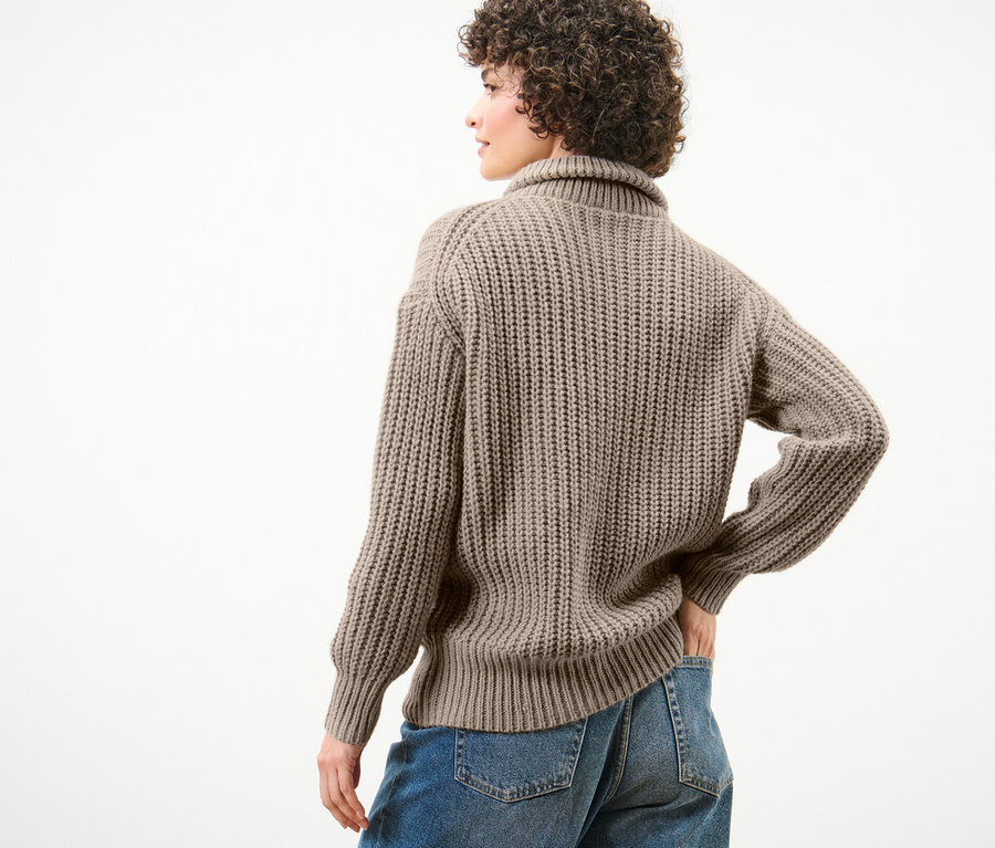 Une femme pose dos à la caméra et porte un pull camionneur taupe en grosse maille et un jean bleu.