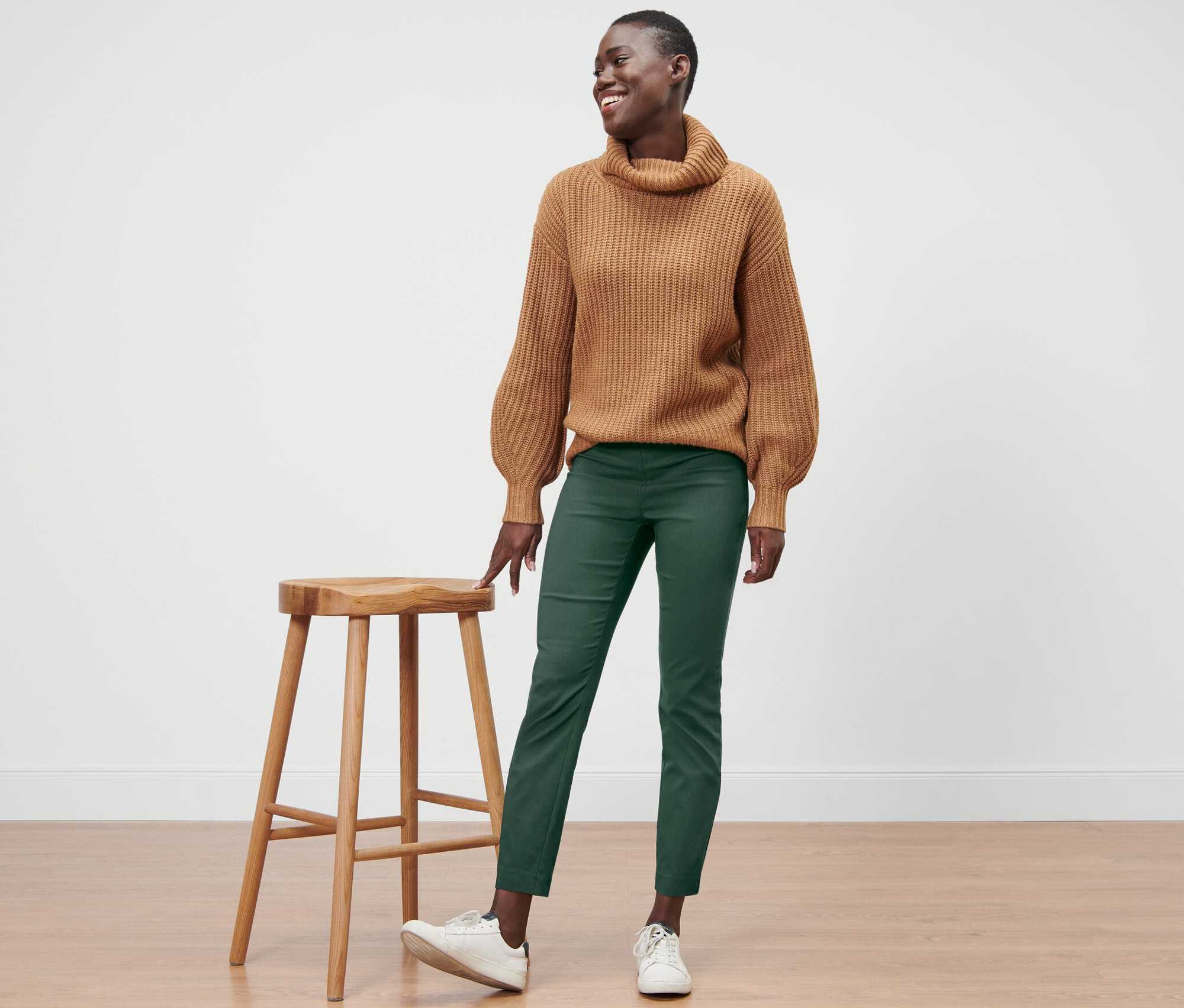 Une femme souriante se tient à côté d'un tabouret en bois, portant un pull marron et un pantalon stretch vert.
