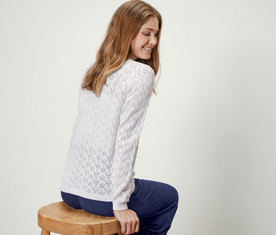 Une femme est assise de côté sur un tabouret et porte une jaquette en maille à motif ajouré, blanc.