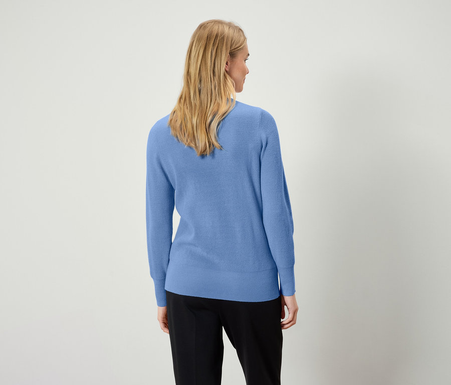 Une femme blonde porte un pull bleu clair et un pantalon noir, vue de dos.