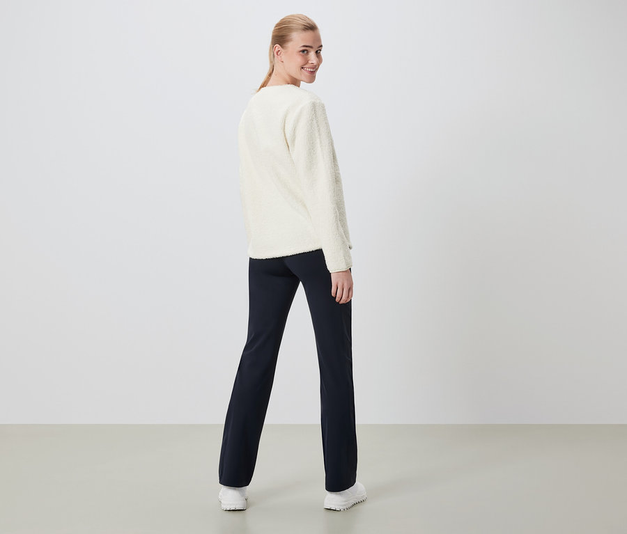 Une femme regarde par-dessus son épaule, portant un pull crème et un pantalon bleu marine.