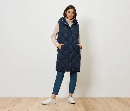 Une femme debout porte un long gilet matelassé bleu marine à capuche, un jean et des baskets.
