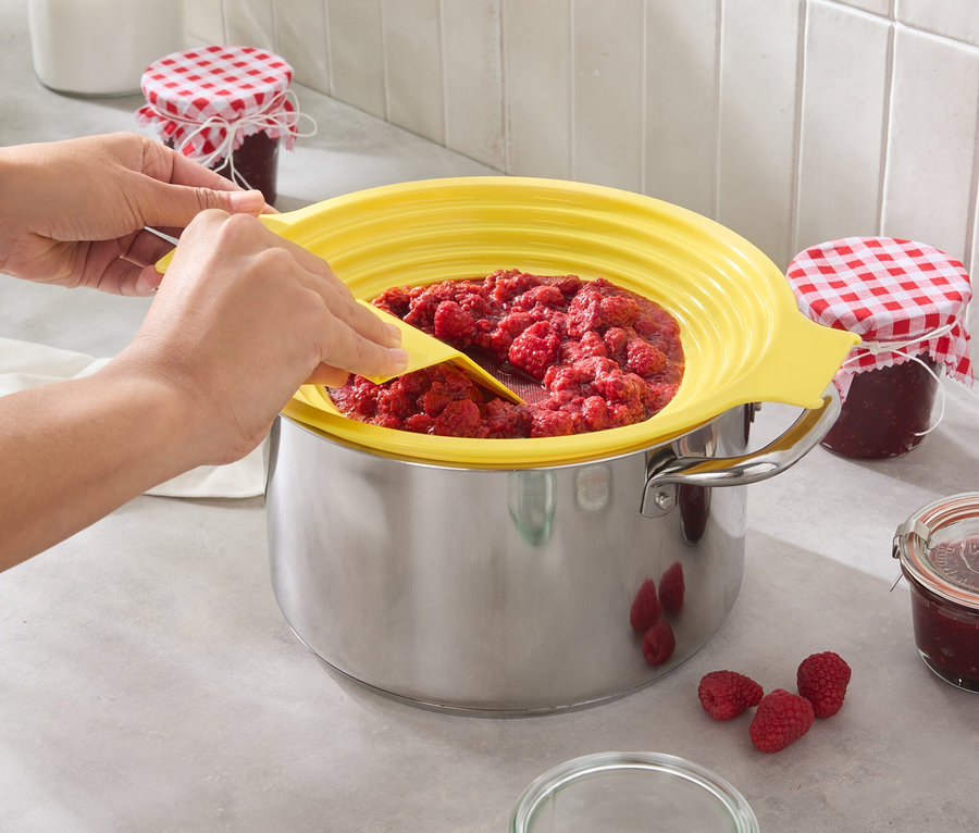 Une personne mélange de la confiture de framboises dans une casserole avec un couvercle anti-éclaboussures jaune. Des framboises et des pots sont posés sur le plan de travail de la cuisine.