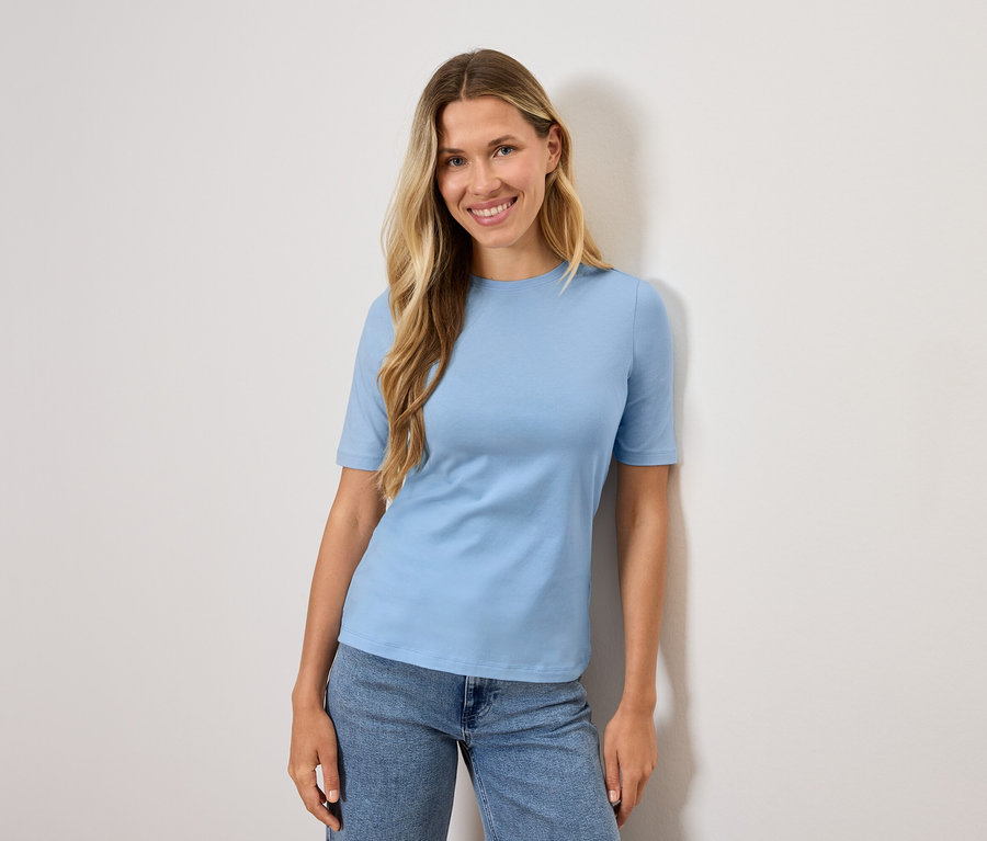 Une femme aux cheveux blonds porte un t-shirt bleu clair et un jean.