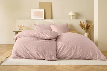 Linge de lit rose sur un lit devant un fond beige.