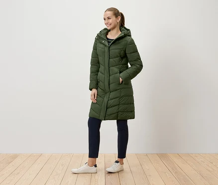 Une femme portant un long manteau matelassé vert avec capuche et un pantalon bleu marine se tenant sur un plancher en bois.