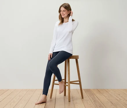 Jeune femme assise sur un tabouret en bois, portant un t-shirt blanc à manches longues et un legging bleu marine.