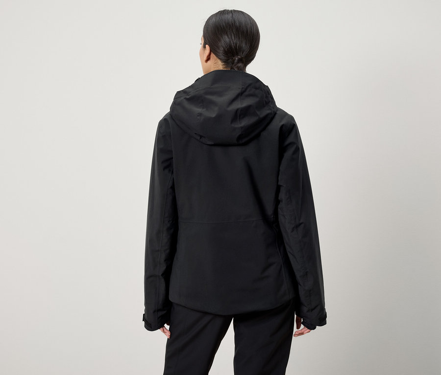 Vue de dos, une femme aux cheveux tirés en queue de cheval porte une veste à capuche noire et un pantalon noir.