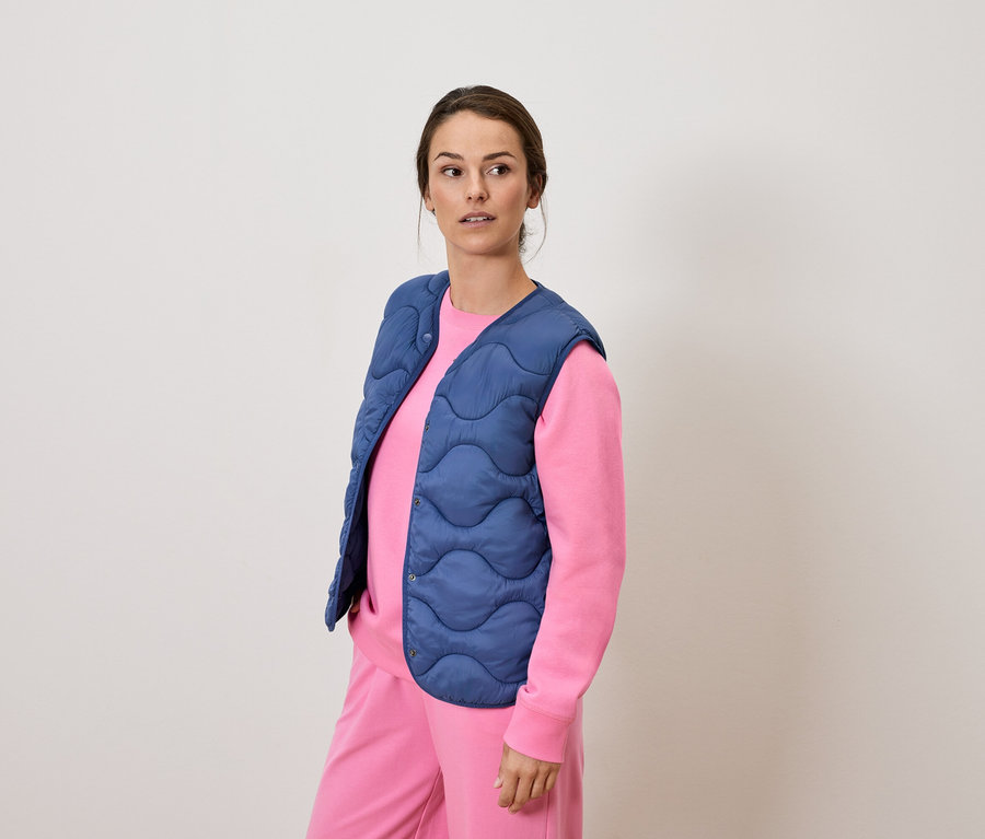 Une femme en survêtement rose et gilet matelassé bleu debout regardant au loin.