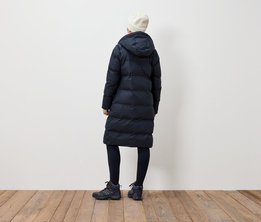 Une femme, vue de dos, porte un manteau en duvet noir, des leggings noirs, des bottes noires et un bonnet blanc.