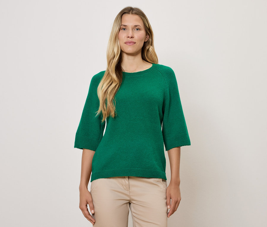 Une femme blonde porte un pull vert à manches trois-quarts et un pantalon beige.