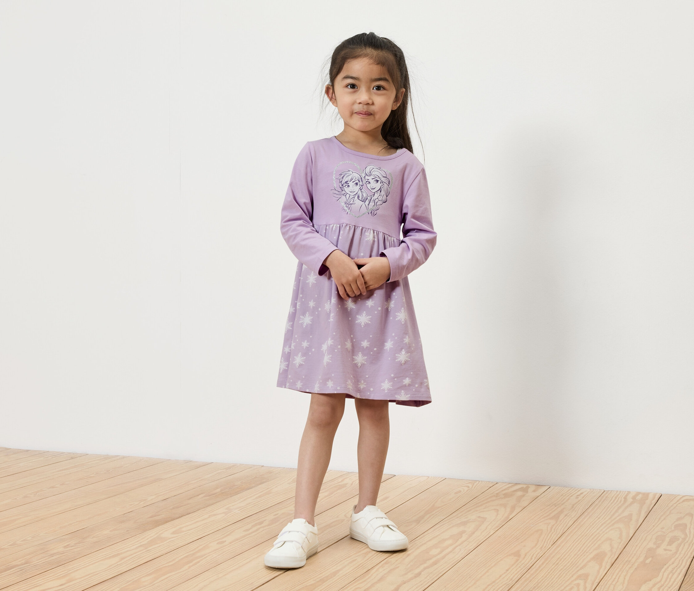 Une fille se tient debout et porte une robe à manches longues violet clair avec un motif Elsa et Anna et des baskets blanches.