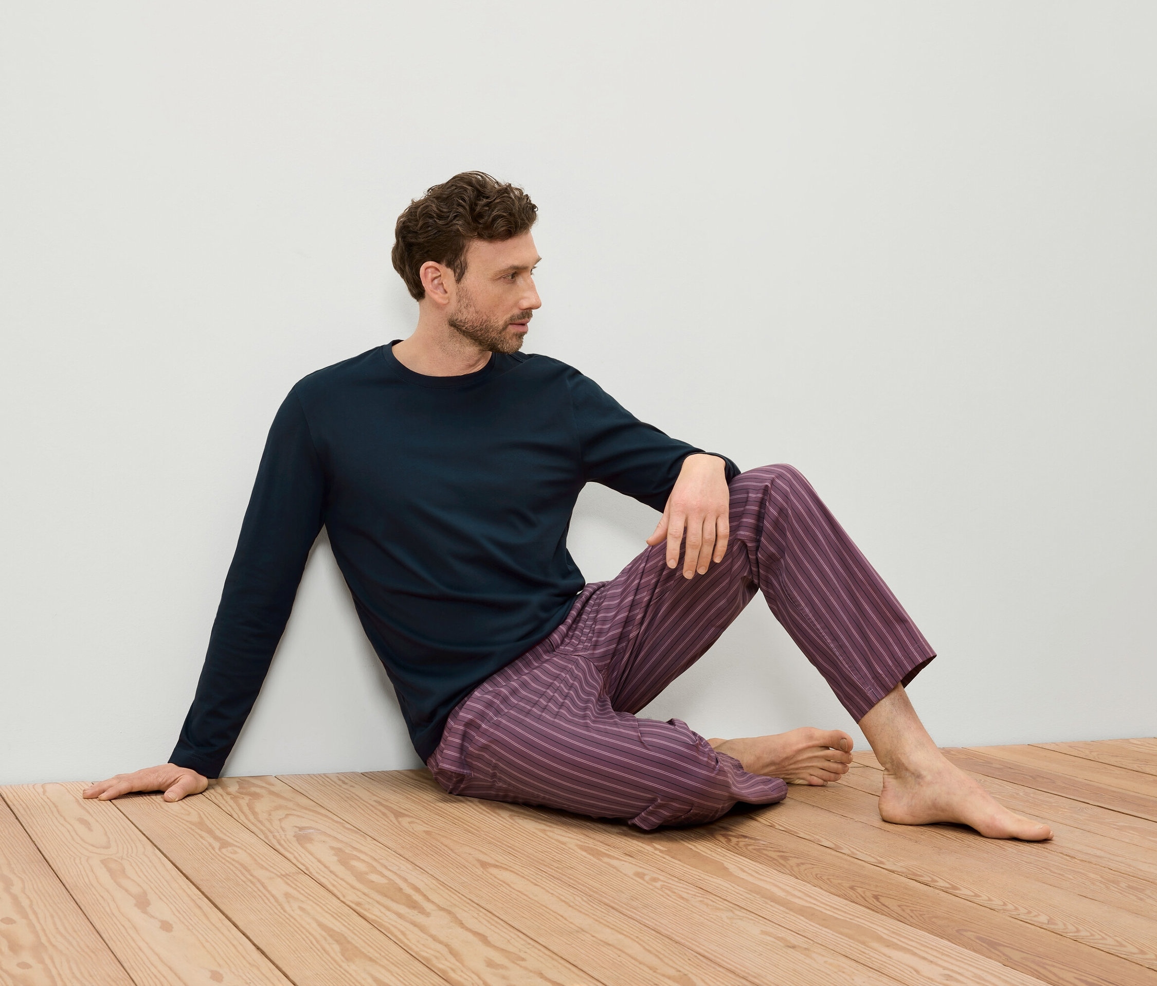 Un homme est assis sur un plancher en bois contre un mur blanc, portant un t-shirt à manches longues bleu foncé et un pantalon rayé violet.