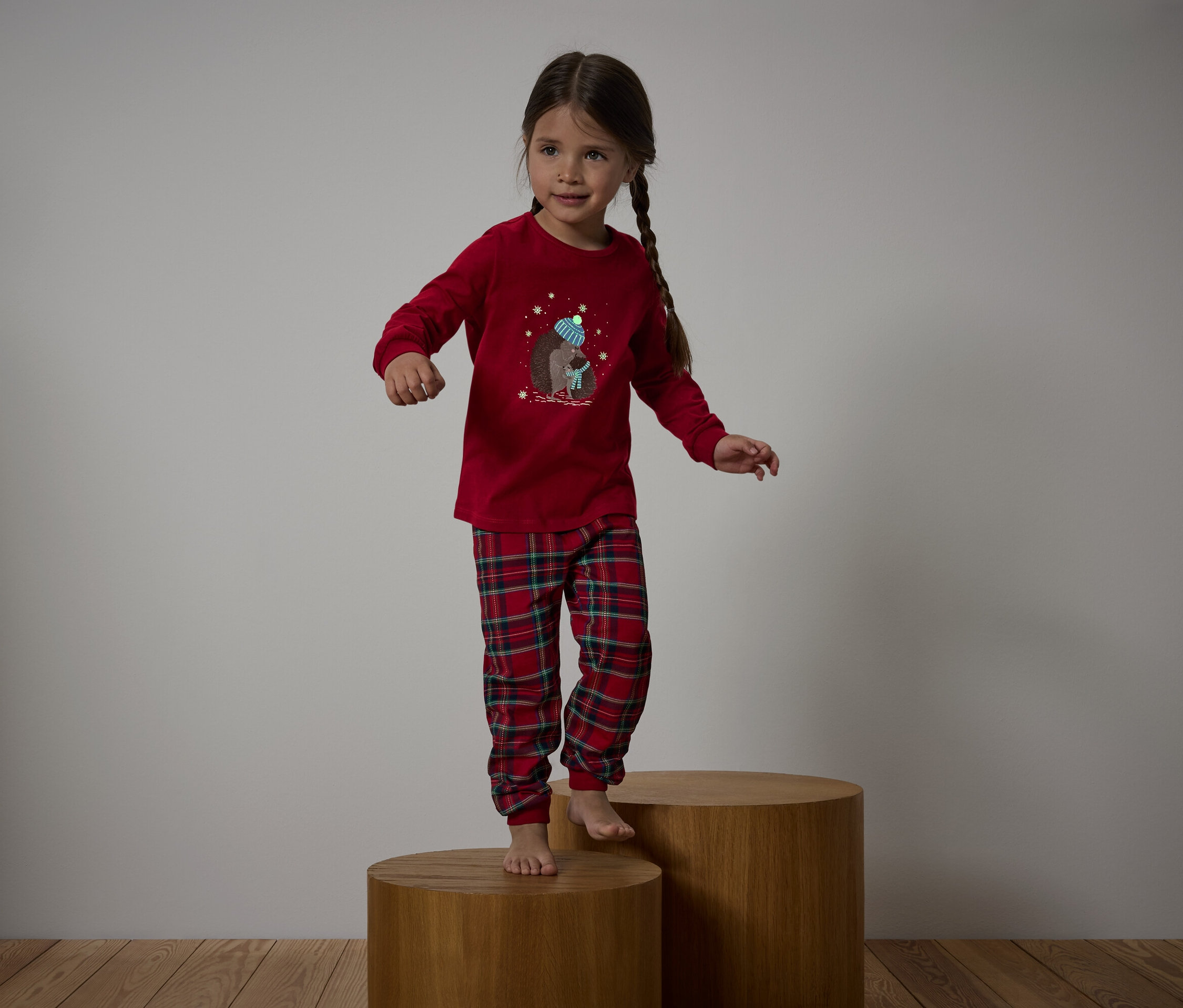 Une fille en pyjama rouge avec un pantalon à carreaux se tient sur des socles en bois.