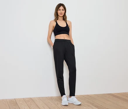 Une femme portant un haut de sport et un pantalon noirs se tient contre un mur blanc.