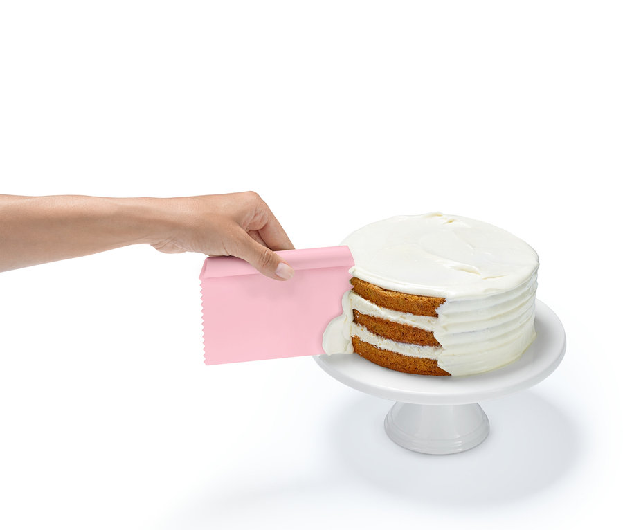 Une main applique du glaçage sur un gâteau à trois étages avec une spatule rose, posé sur un socle blanc.