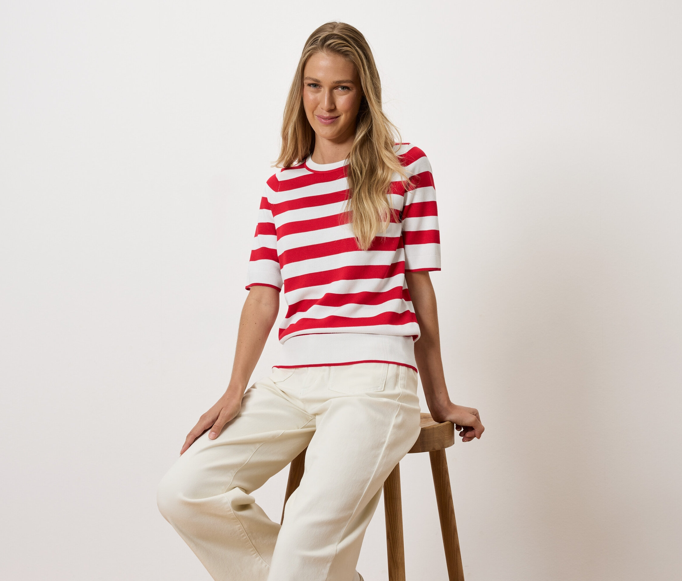 Une femme blonde est assise sur un tabouret en bois, portant un t-shirt rayé rouge et blanc et un pantalon blanc.