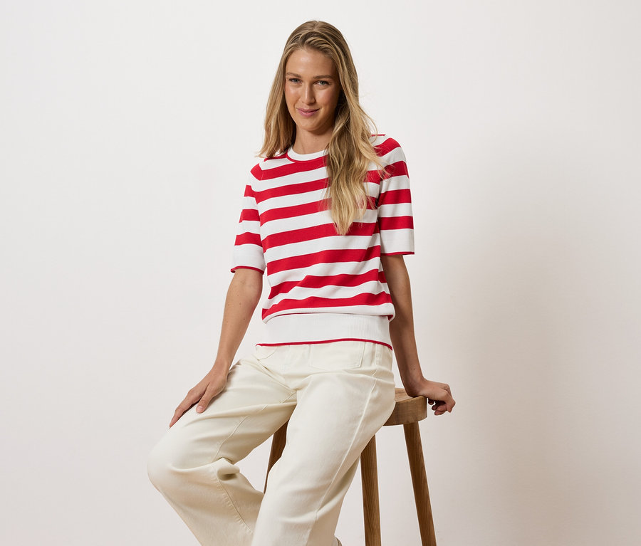 Une femme blonde est assise sur un tabouret en bois, portant un t-shirt rayé rouge et blanc et un pantalon blanc.