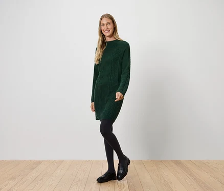 Une femme portant une robe verte en tricot, des collants noirs et des chaussures noires pose sur un plancher en bois devant un mur blanc.