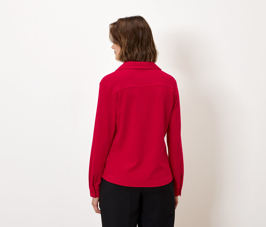 De dos, une femme aux cheveux bruns porte une chemise rouge à manches longues et un pantalon noir.