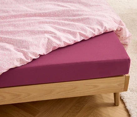 Gros plan d'un lit en bois avec un drap-housse en jersey de taille standard couleur baies et du linge de lit rose.