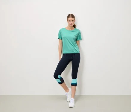 Une femme se tient devant un mur blanc, portant un t-shirt turquoise, un legging bleu marine avec des détails turquoise et des baskets blanches.