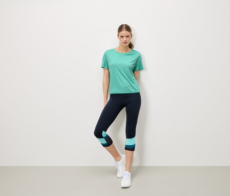 Une femme se tient devant un mur blanc, portant un t-shirt turquoise, un legging bleu marine avec des détails turquoise et des baskets blanches.