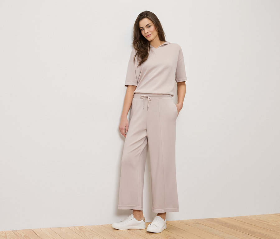 Une femme se tient devant un mur blanc, vêtue d'une tenue beige et de baskets blanches.