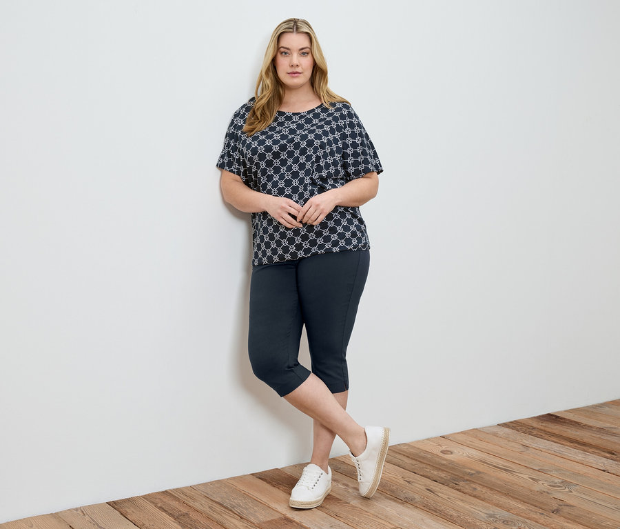 Une femme portant un haut à motifs, un pantalon capri bleu et des chaussures blanches est appuyée contre un mur blanc.