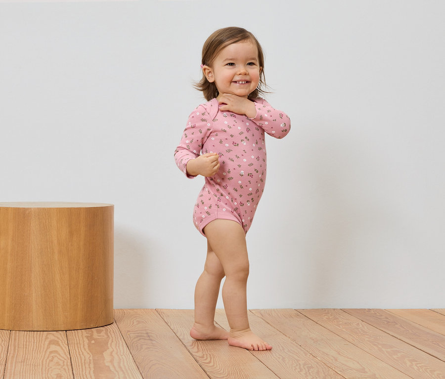 Un bébé souriant portant un body rose à fleurs se tient à côté d'un tabouret en bois sur un plancher en bois.