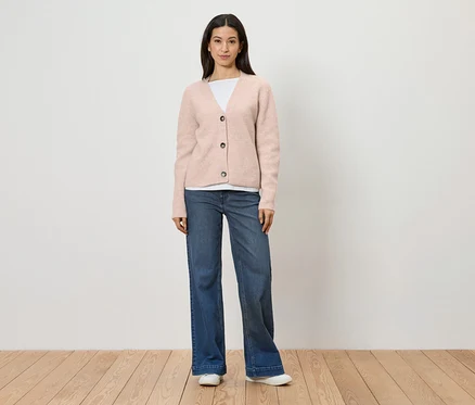 Une femme portant un cardigan rose, un t-shirt blanc, un jean bleu et des chaussures blanches se tient devant un mur blanc.
