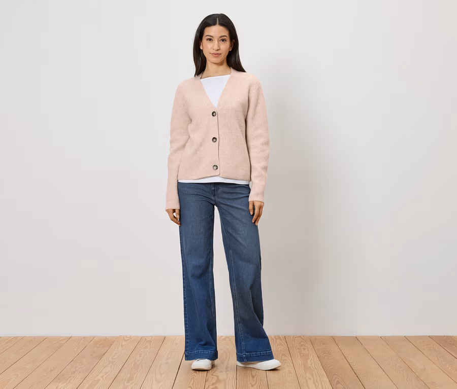 Une femme portant un cardigan rose, un t-shirt blanc, un jean bleu et des chaussures blanches se tient devant un mur blanc.