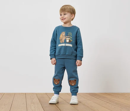 Un garçon aux cheveux blonds porte un pull bleu avec un motif d'ours et de raton laveur et un pantalon bleu avec des patchs d'ours sur les genoux.