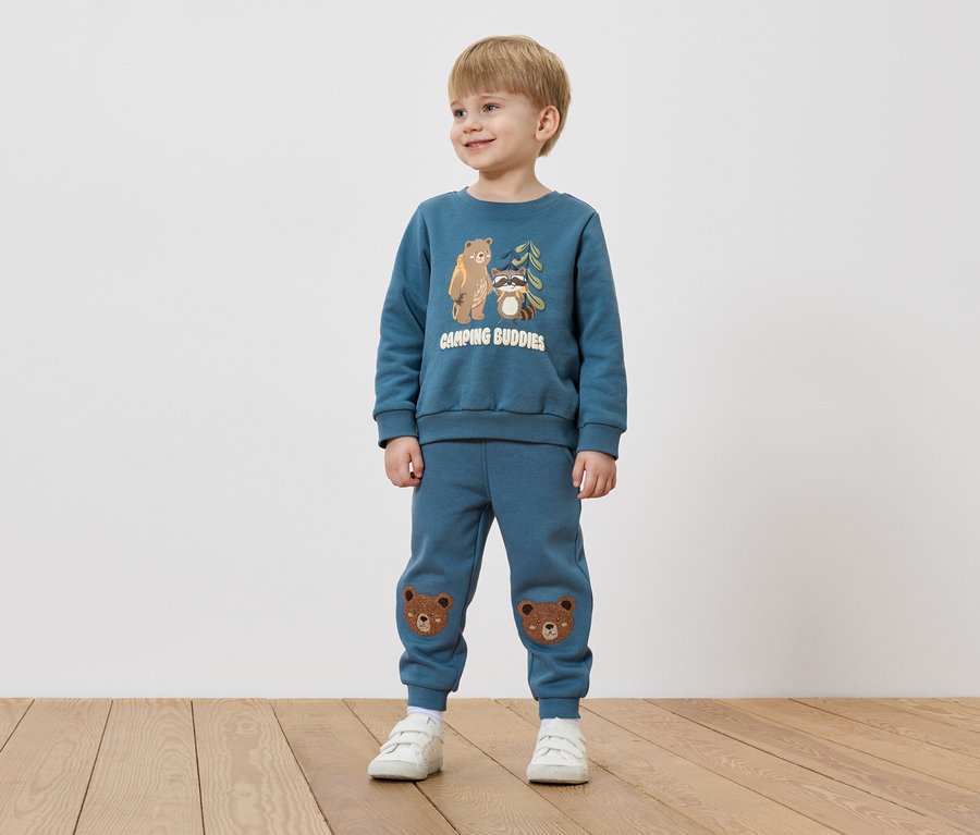 Un garçon aux cheveux blonds porte un pull bleu avec un motif d'ours et de raton laveur et un pantalon bleu avec des patchs d'ours sur les genoux.
