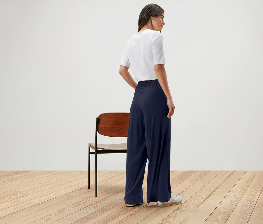 Femme debout, dos à la caméra, portant un t-shirt blanc et un pantalon ample bleu marine.