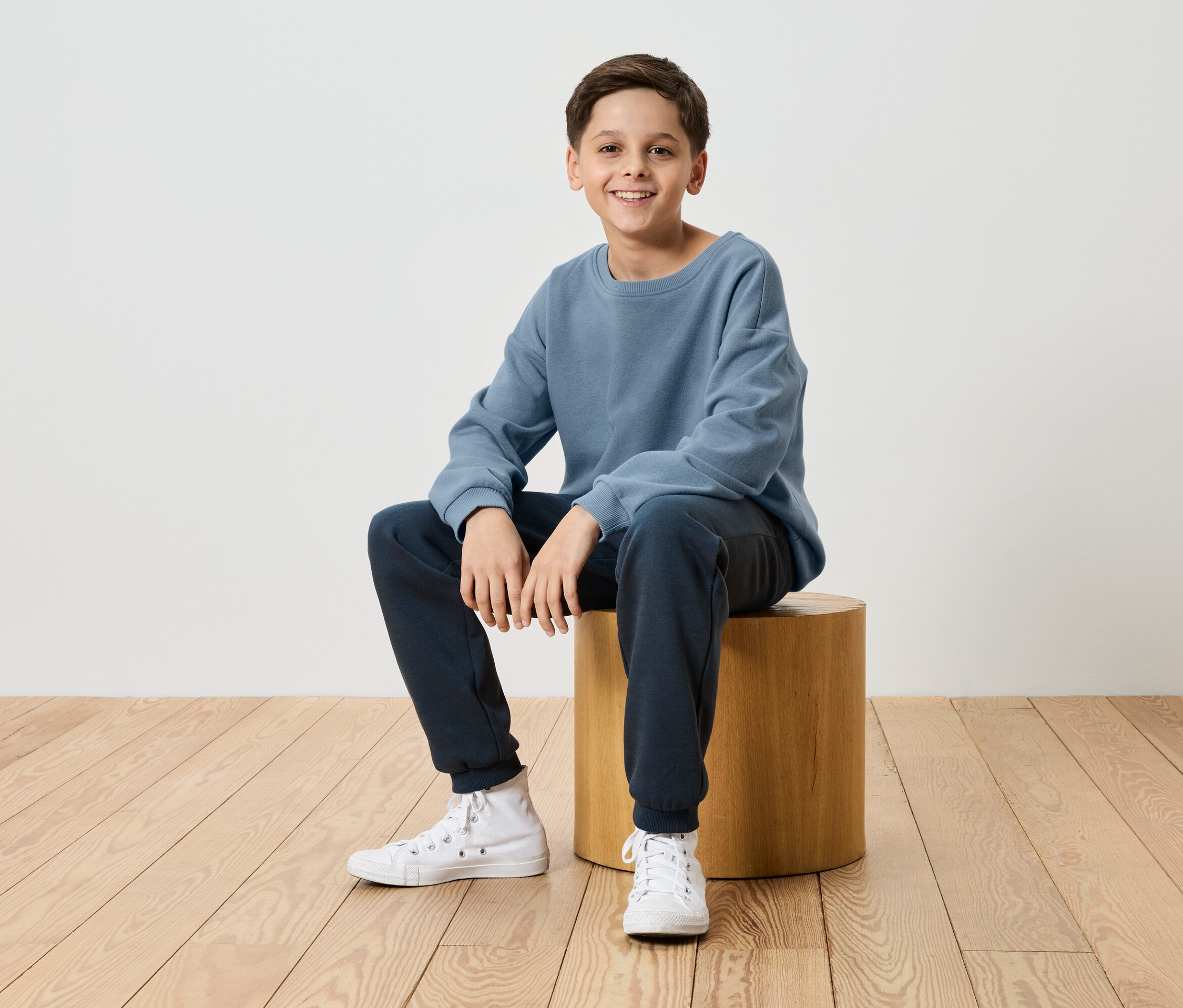 Un garçon souriant est assis sur un tabouret en bois, vêtu d'un pull bleu, d'un pantalon de jogging bleu marine et de baskets blanches.