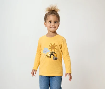 Une fillette aux cheveux bouclés porte un t-shirt jaune à manches longues avec un nuage, un soleil et un arc-en-ciel, et un jean bleu.