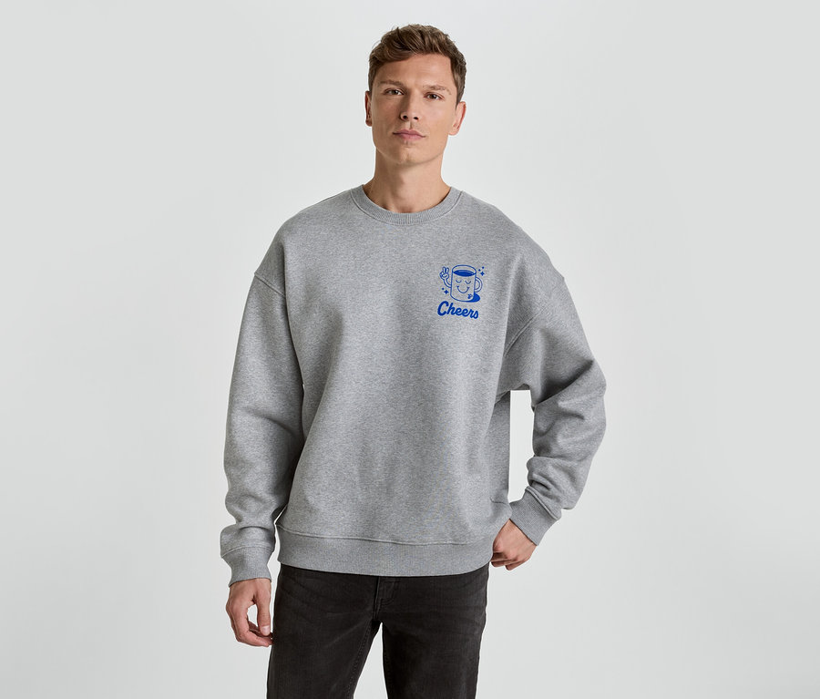Un homme porte un sweat-shirt gris avec l'inscription Cheers et une tasse en bleu.