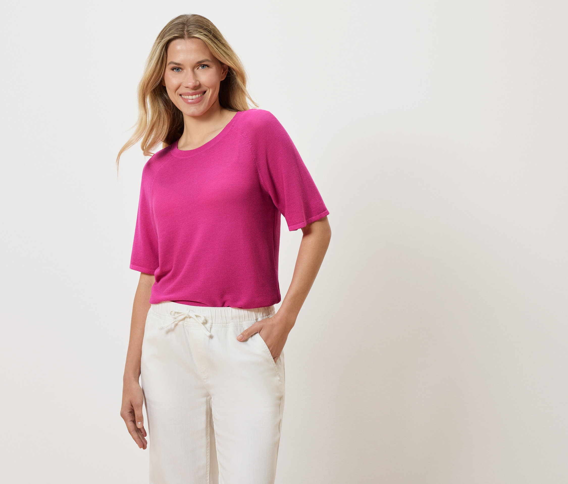 Une femme aux cheveux blonds ondulés porte un t-shirt rose et un pantalon blanc, avec une main dans sa poche.