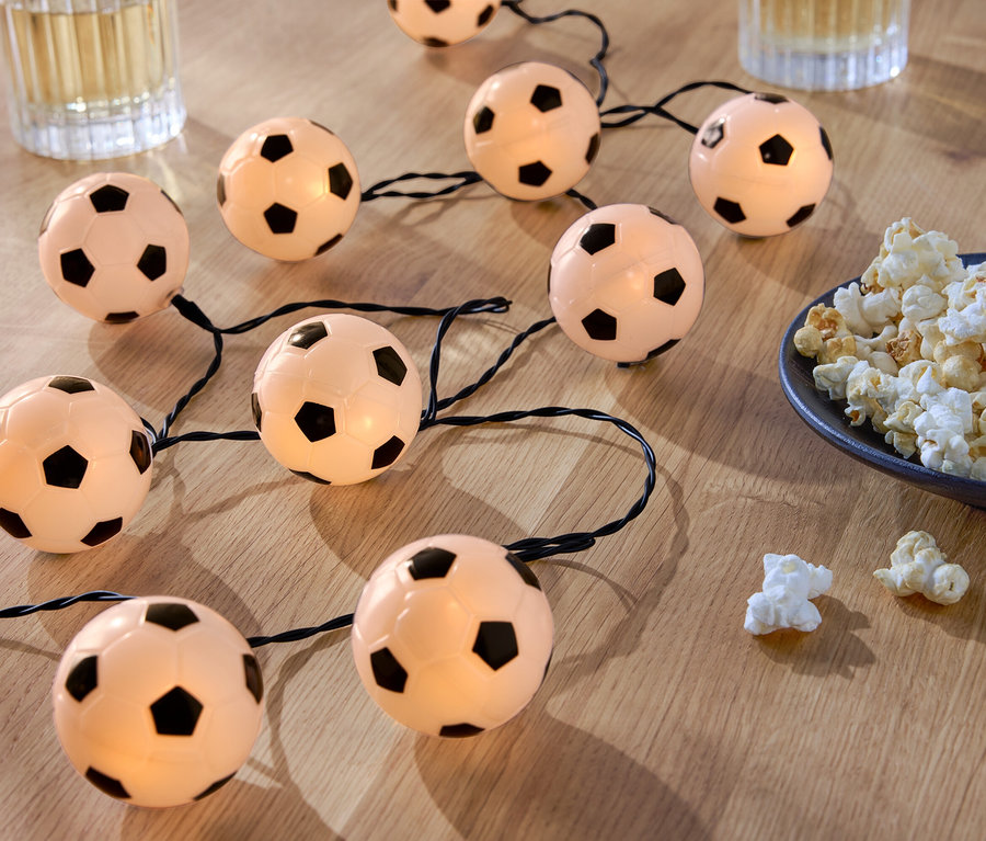 Une guirlande lumineuse en forme de ballon de football repose sur une table en bois à côté d'un bol de pop-corn et de deux verres de boissons.