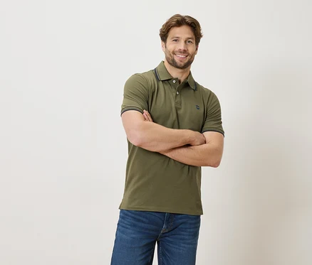 Un homme aux bras croisés porte un polo vert et un jean sur fond blanc.