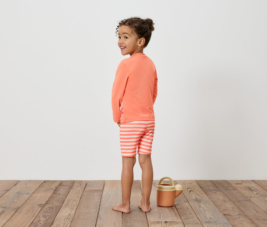 Fillette portant un haut orange et un short rayé, à côté d'un arrosoir sur un plancher en bois.