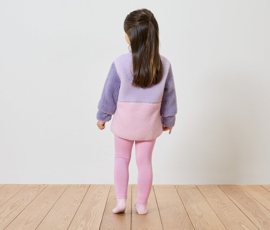 Vue arrière d'une fillette aux longs cheveux bruns portant un pull en peluche lavande et rose à blocs de couleurs et un legging rose.