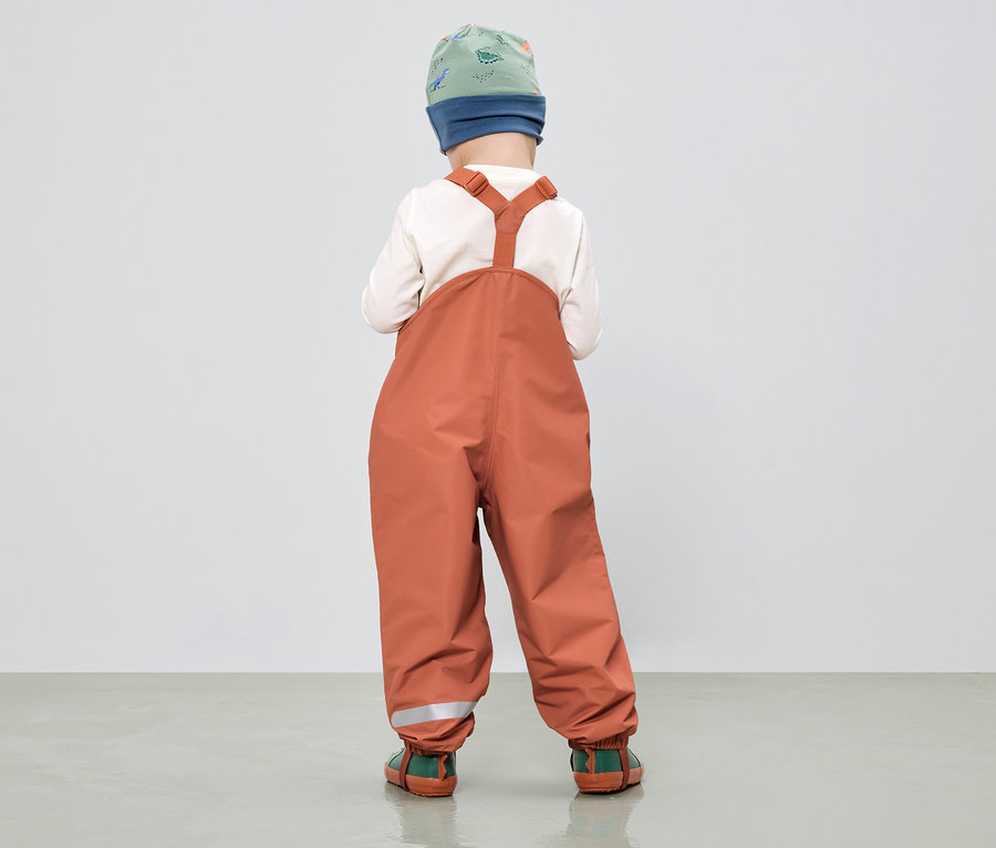Enfant vu de dos portant une salopette de pluie orange, un bonnet à motifs vert et bleu et un haut crème.