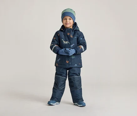 Un garçon en vêtements d'hiver bleu marine à motifs de dinosaures, bonnet et gants, se tient devant un fond clair.
