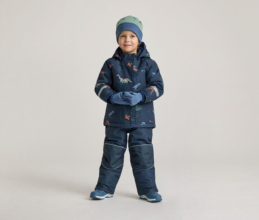 Un garçon en vêtements d'hiver bleu marine à motifs de dinosaures, bonnet et gants, se tient devant un fond clair.